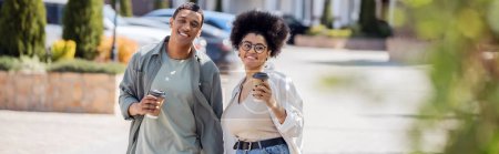 pareja afroamericana positiva con café para llevar caminando por la calle urbana borrosa, pancarta