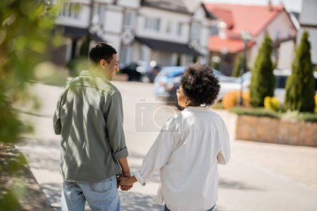 Foto de Vista lateral de alegre pareja afroamericana hablando y tomándose de la mano y caminando por la calle urbana - Imagen libre de derechos