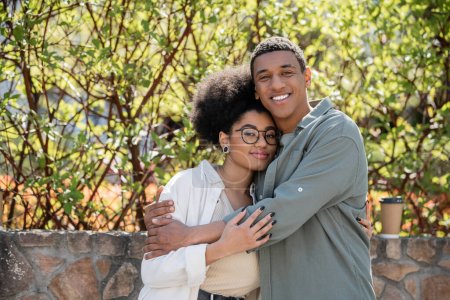 smiling african american man hugging girlfriend and looking at camera near coffee to go outdoors