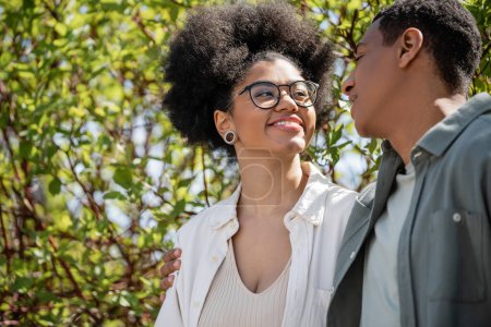 smiling african american woman in eyeglasses looking at boyfriend on urban street in summer