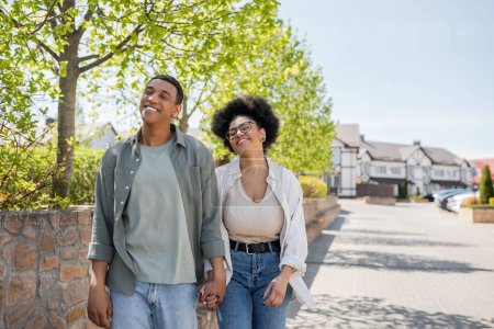 positive african american couple holding hands while walking on urban street in summer