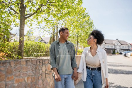 smiling african american couple holding hands and talking while walking on urban street in summer