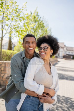 sonriente afroamericano hombre abrazando novia en gafas y mirando a la cámara en la calle urbana