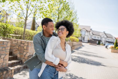 glücklicher afrikanisch-amerikanischer Mann umarmt Freundin in Brille auf urbaner Straße im Sommer