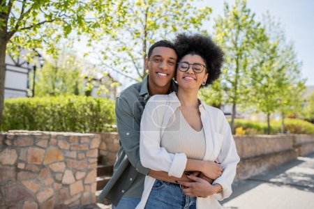 african american man embracing girlfriend in eyeglasses while standing together outdoors in summer