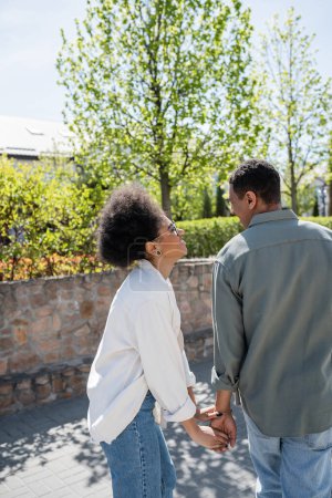 positive african american woman in eyeglasses holding hand of boyfriend on street in summer