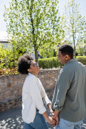 glückliches afrikanisch-amerikanisches Liebespaar hält Händchen, während es im Sommer auf der Straße spaziert