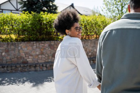 Foto de Sonriente afroamericana mujer en camisa y gafas mirando novio en la calle urbana - Imagen libre de derechos