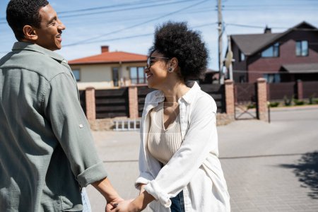 Photo for Side view of smiling african american couple holding hands on urban street with houses at background - Royalty Free Image