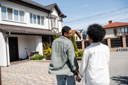 lächelnder afrikanisch-amerikanischer Mann hält Hand seiner Freundin in der Nähe des verschwommenen Hauses im Hintergrund