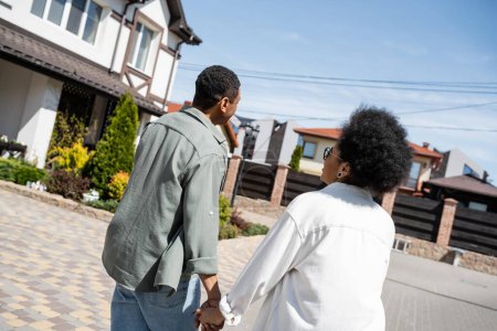 joyful african american couple holding hands near blurred house on street in summer