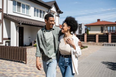 smiling man hugging african american girlfriend while walking on sidewalk near houses on street