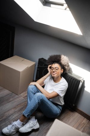 Hochwinkelaufnahme einer lächelnden afrikanisch-amerikanischen Frau, die in der Nähe von Kartons auf dem Dachboden in einem Haus sitzt