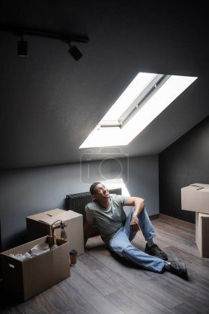 cheerful african american man sitting in sunlight near carton boxes on attic in new house