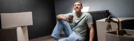 Téléchargez les photos : Homme afro-américain joyeux tenant du café pour aller près des boîtes en carton dans une nouvelle maison, bannière - en image libre de droit