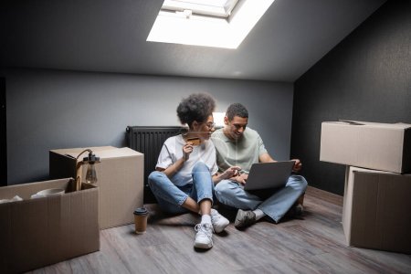 sonriente africano americano pareja usando portátil y tarjeta de crédito cerca de cajas en ático en nueva casa