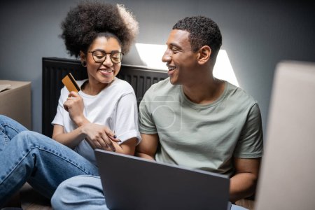 Téléchargez les photos : Joyeux couple afro-américain en utilisant un ordinateur portable et une carte de crédit près de boîtes en carton dans une nouvelle maison - en image libre de droit