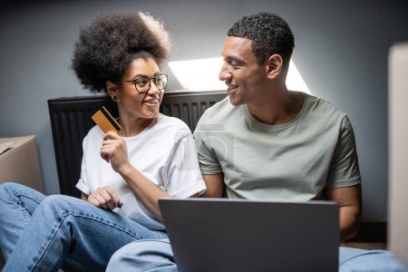 Téléchargez les photos : Souriante femme afro-américaine tenant carte de crédit près de petit ami avec ordinateur portable dans une nouvelle maison - en image libre de droit