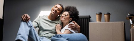 pareja afroamericana positiva abrazándose cerca del café en caja de cartón en ático en casa nueva, pancarta