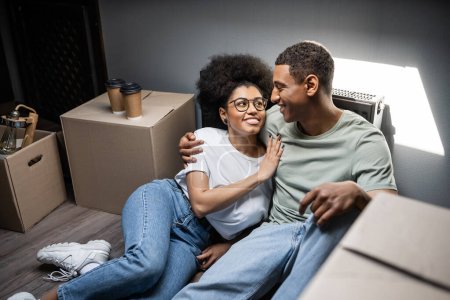 cheerful african american woman hugging boyfriend near coffee to go and boxes in new house