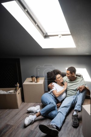 Sonriente pareja afroamericana abrazándose cerca de cajas de café y cartón en el ático en una casa nueva