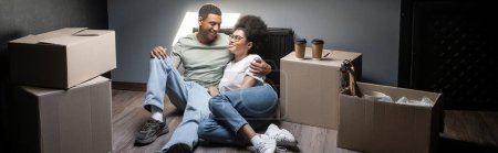 smiling african american couple hugging near carton boxes and takeaway coffee in new house, banner