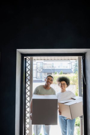 Lächelndes afrikanisch-amerikanisches Paar mit Kartons in der Hand und Blick in die Kamera in der Nähe der Tür in einem neuen Haus