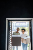 smiling african american couple holding carton boxes and looking at camera near door in new house mug #667987002