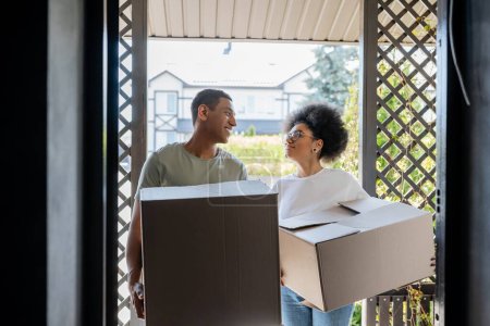 Téléchargez les photos : Sourire romantique couple afro-américain tenant des boîtes en carton près de la porte de la nouvelle maison - en image libre de droit