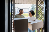 smiling romantic african american couple holding carton boxes near door of new house Poster #667987010