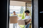 happy african american man holding cardboard box near girlfriend with package and door of new house mug #667987026