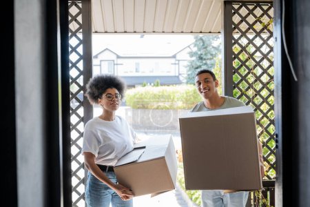Téléchargez les photos : Joyeux couple afro-américain avec des paquets regardant la caméra près de la porte de la nouvelle maison - en image libre de droit