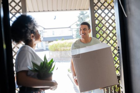 souriant homme afro-américain tenant boîte de carton près de petite amie avec plante dans la porte de la nouvelle maison