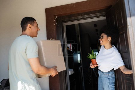 heureux couple afro-américain tenant plante et boîte en carton près de la porte de la nouvelle maison