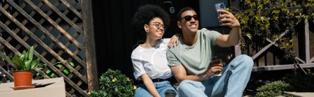 couple afro-américain positif avec prise de café selfie près de boîtes en carton et près de la maison, bannière