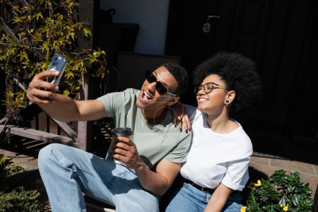 alegre africano americano pareja con café tomando selfie en smartphone cerca de nueva casa