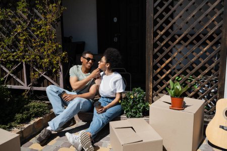 pareja afroamericana positiva con café para ir sentado cerca de cajas de cartón y casa nueva