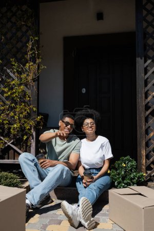Téléchargez les photos : Souriant homme afro-américain avec café pointant vers des boîtes près de petite amie sur le porche de la nouvelle maison - en image libre de droit