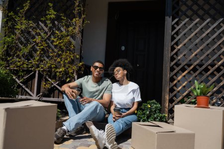 Téléchargez les photos : Heureux couple afro-américain tenant du café et assis près des paquets et de la nouvelle maison - en image libre de droit