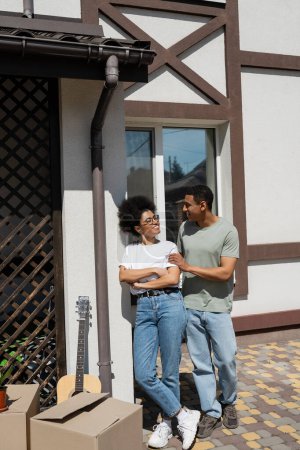 sonriente pareja afroamericana de pie cerca de cajas de cartón y nueva casa al aire libre