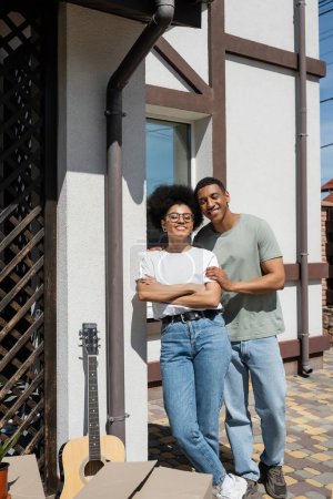 lächelnder afrikanisch-amerikanischer Mann umarmt Freundin in der Nähe von Akustikgitarre und Boxen in der Nähe von neuem Haus