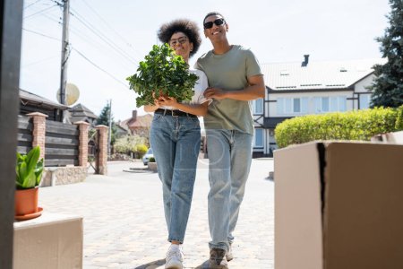 Photo for Cheerful african american couple holding houseplant near cardboard boxes during relocation outdoors - Royalty Free Image