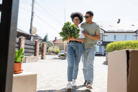pareja afroamericana positiva sosteniendo planta de interior cerca de cajas de cartón durante la reubicación al aire libre
