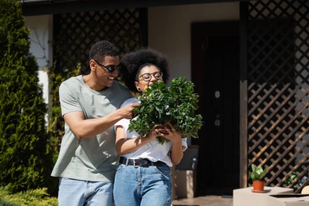 heureux couple afro-américain tenant plante d'intérieur près de boîtes en carton et nouvelle maison à l'extérieur