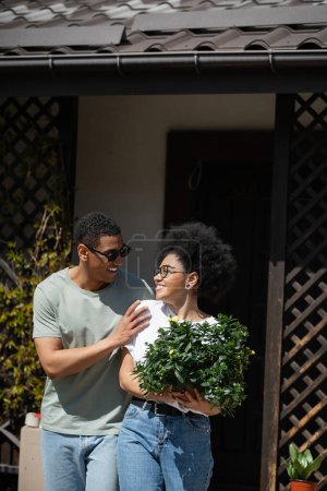 sonriente africano americano hombre en gafas de sol abrazando novia con houseplant cerca de nueva casa