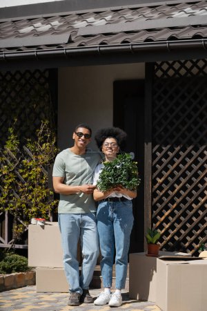 feliz africano americano pareja holding houseplant cerca paquetes y nuevo casa al aire libre
