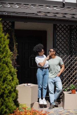 sonriente afroamericana mujer tocando novio mientras de pie cerca de paquetes y nueva casa