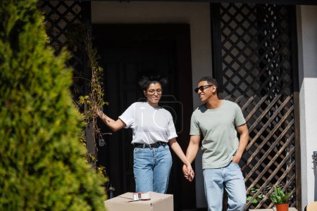 sonriente africano americano pareja cogido de la mano cerca paquete y nuevo casa en fondo