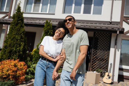 smiling african american woman in eyeglasses hugging boyfriend near new house at background