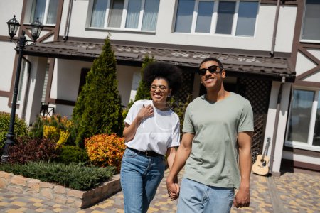 pareja afro-americana romántica positiva cogida de la mano mientras está de pie cerca de nueva casa